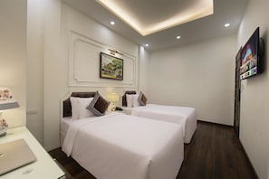 A hotel room with two beds, a desk with a laptop, a bedside table with a lamp, and a painting on the wall. - Bastra Classy Hotel (Hoan Kiem Ward)