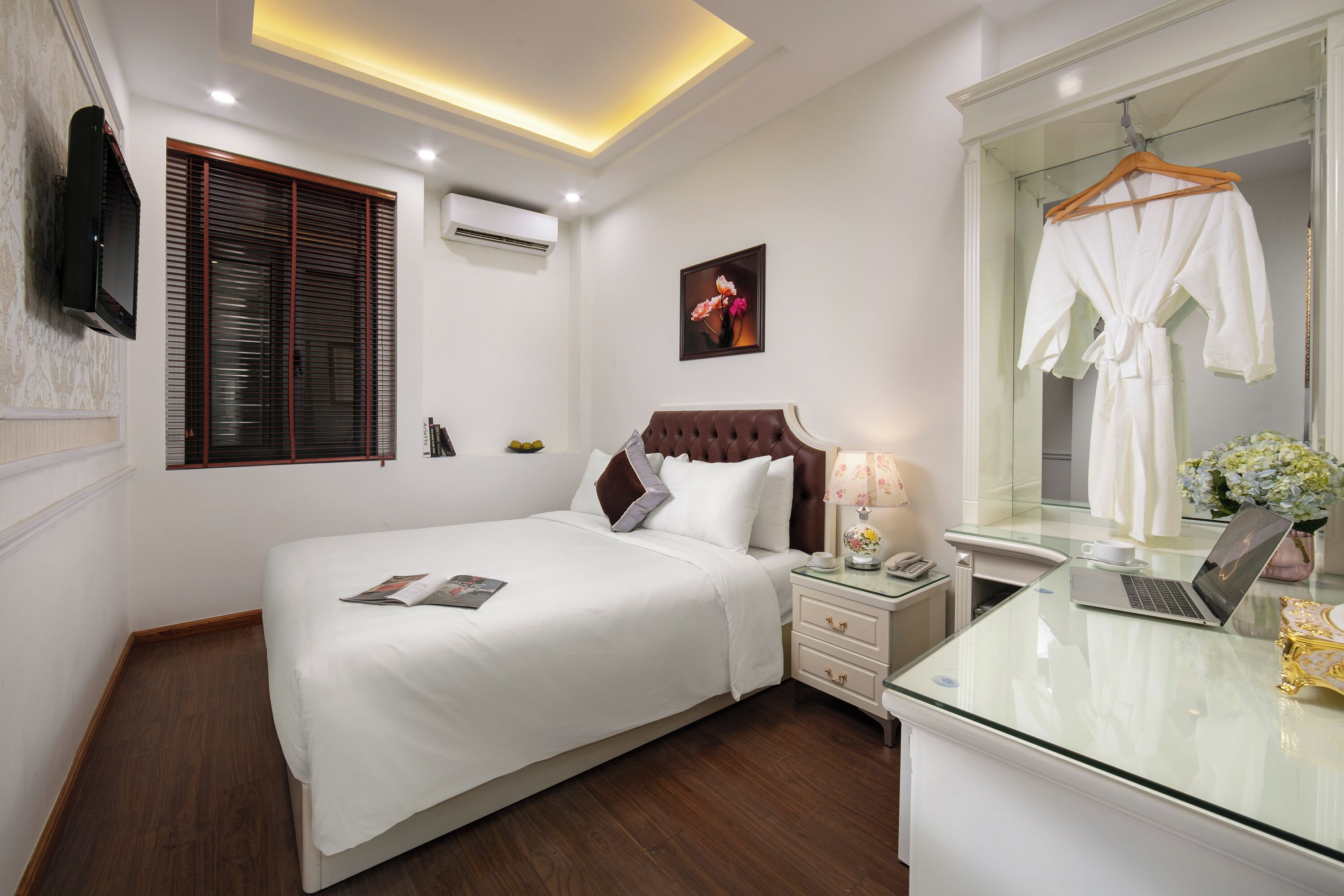 A neatly arranged hotel room with a bed, bedside table, lamp, laptop, and a mirror with a robe hanging.