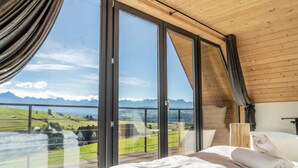 Ein Schlafzimmer mit großem Fenster, einem Bett und Blick auf eine grüne Landschaft.