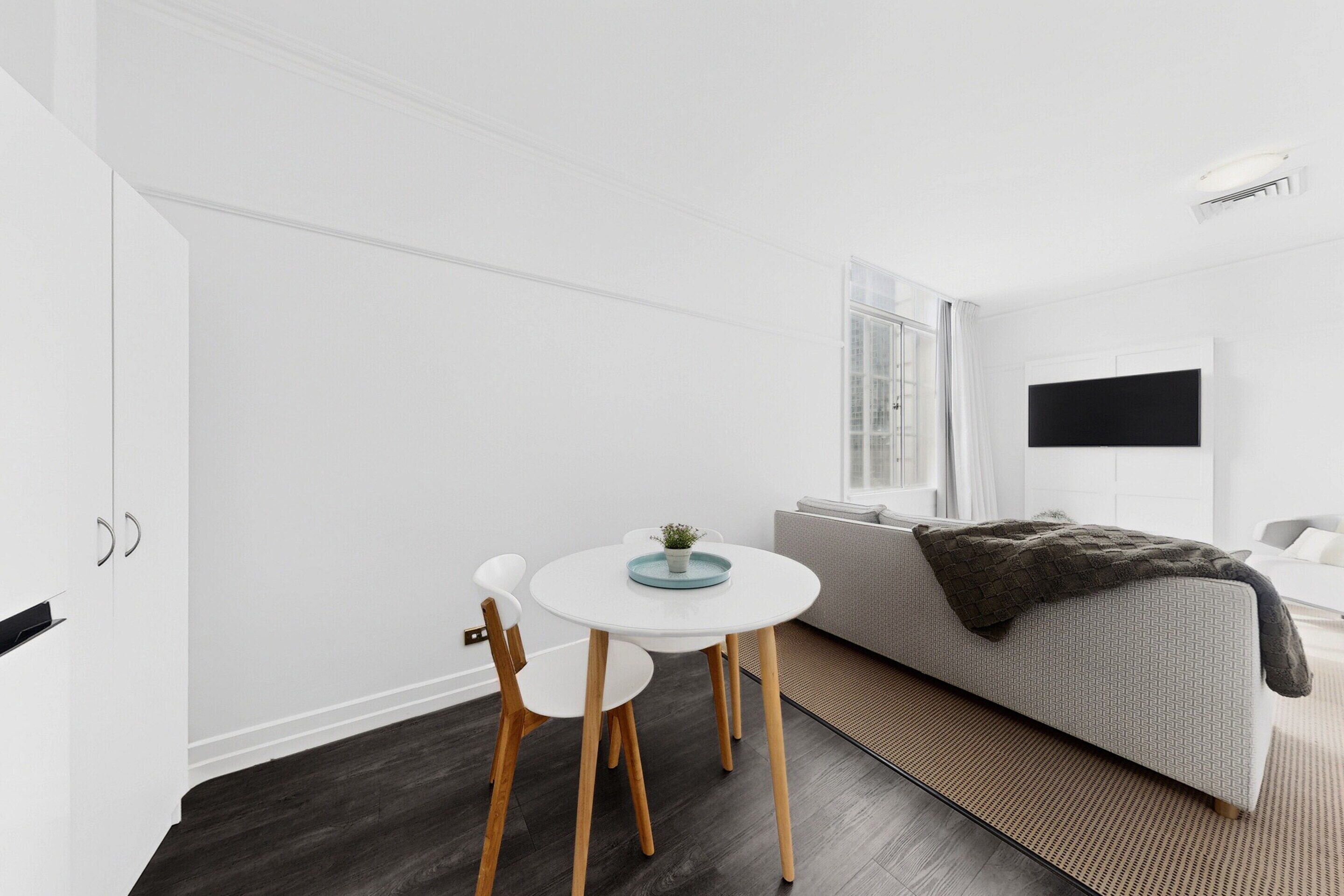 Ein modernes Zimmer mit einem weißen Esstisch, zwei Stühlen, einem grauen Sofa und einem an der Wand befestigten Fernseher.