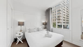 A hotel room with a large bed, a nightstand with a lamp, a window with a view of buildings, and a mirror on the wall.