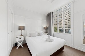 A hotel room with a large bed, a nightstand with a lamp, a window with a view of buildings, and a mirror on the wall. - Skyline 1-bed Retreat in Brisbane City (Brisbane)