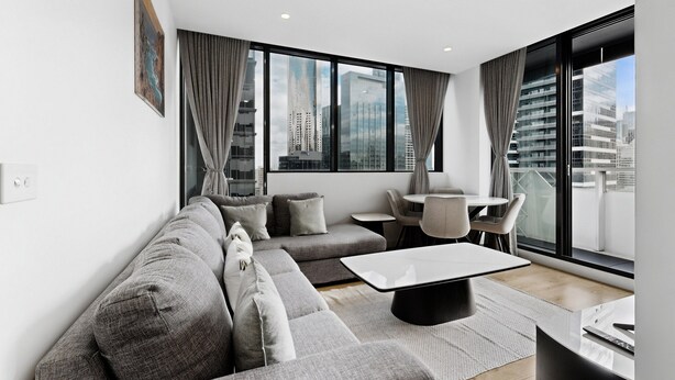 A modern living room with a sectional sofa, a coffee table, and a dining area with chairs and a table. Large windows offer a city view. - Elevated 2-bed Stay Steps From Crown (Melbourne)