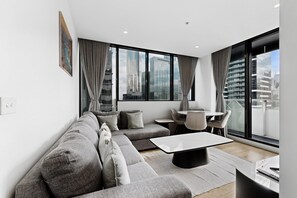 A modern living room with a sectional sofa, a coffee table, and a dining area with chairs and a table. Large windows offer a city view. - Elevated 2-bed Stay Steps From Crown (Melbourne)