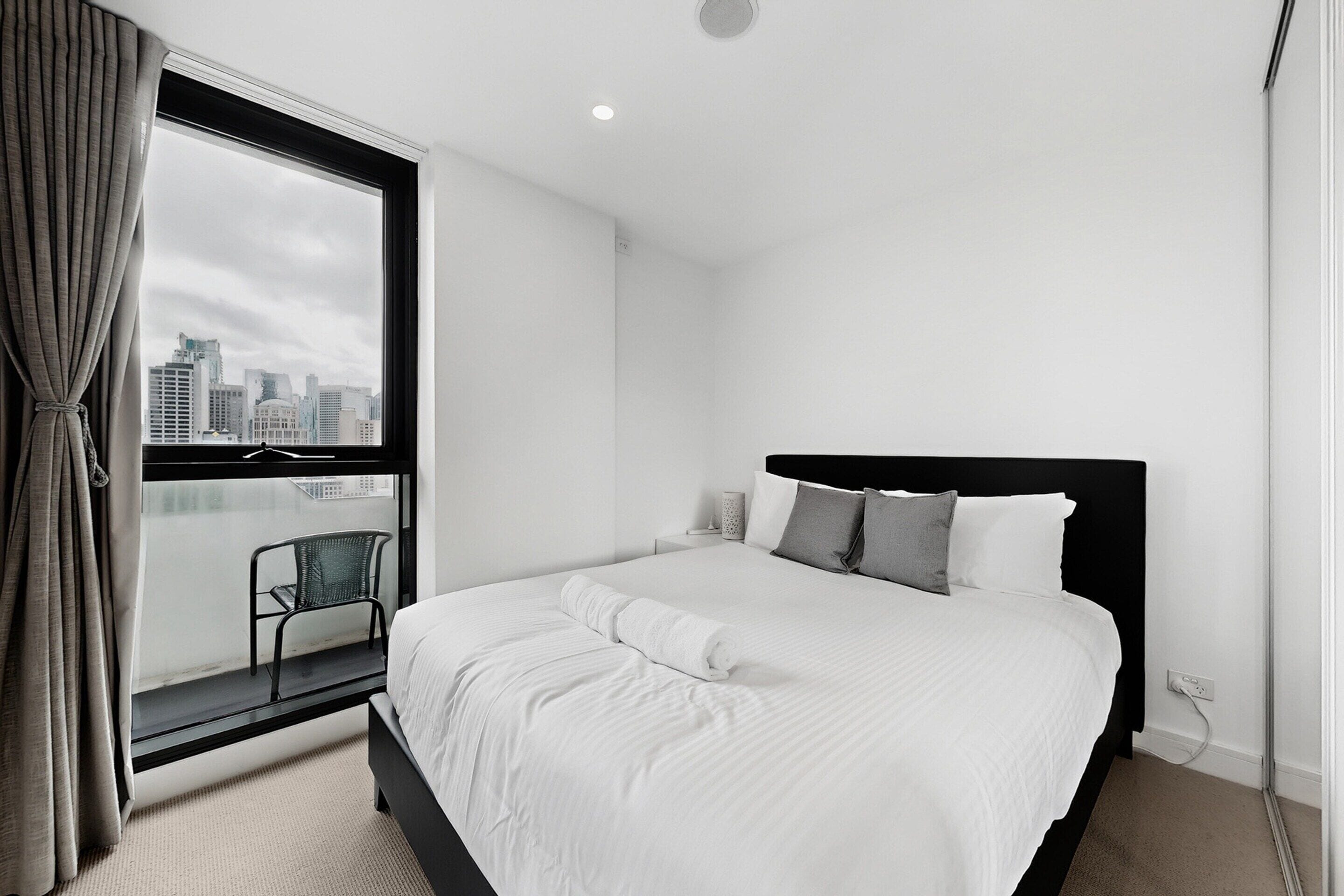 A modern hotel room with a large bed, a window with a city view, a chair by the window, and a black headboard.
