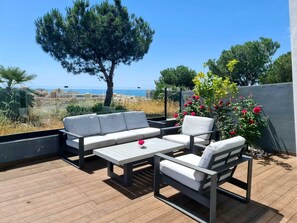 An outdoor seating area with a view of the sea, a large tree, and a flower bush.
