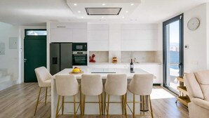 Uma cozinha moderna com armários brancos, uma ilha central com banquetas, geladeira, forno e uma garrafa de vinho no balcão.