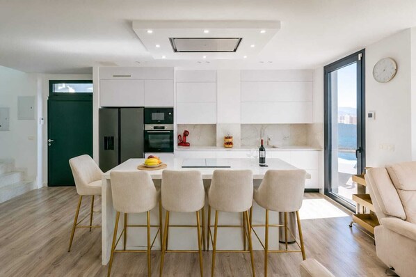 Una cocina moderna con armarios blancos, una isla central con taburetes de bar, un refrigerador, un horno y una botella de vino sobre la encimera.