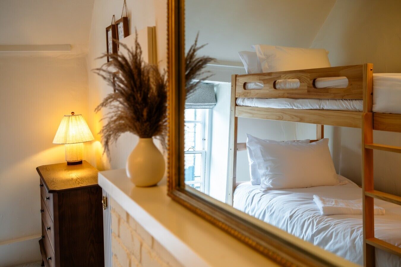 A bunk bed with white bedding, a bedside lamp, a vase with dried plants, and a mirror reflecting the room.