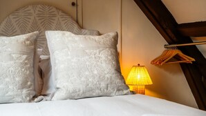 Cama com roupa de cama branca e duas almofadas decorativas, luminária de cabeceira e cabeceira de madeira.