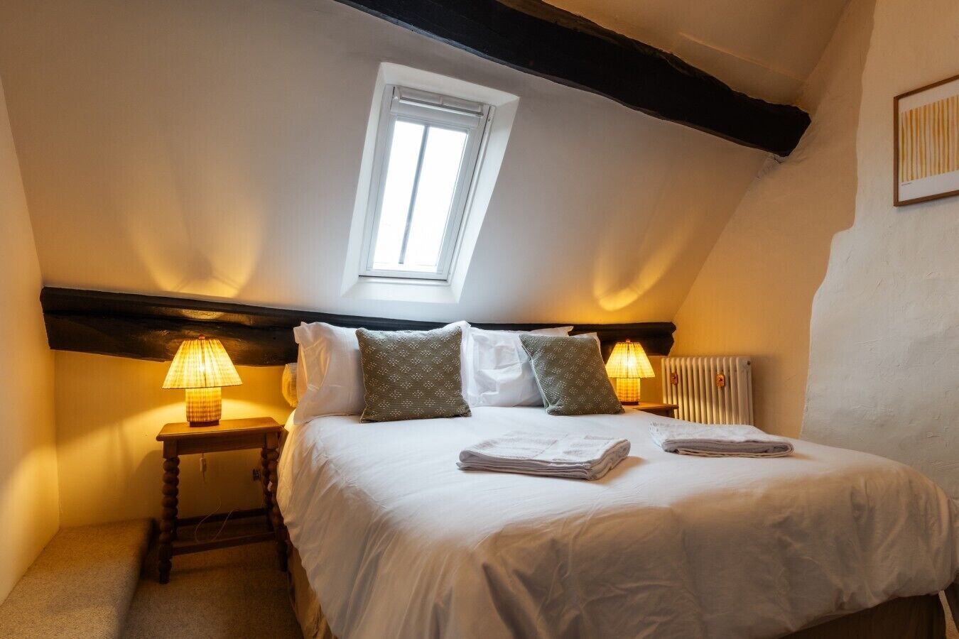 A bedroom with a sloped ceiling, a skylight, a bed with white linens, two bedside lamps, and a radiator.