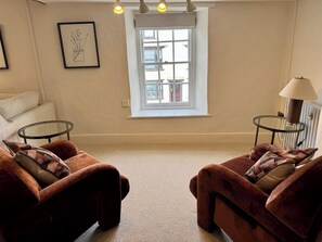 Ein Wohnzimmer mit zwei braunen Sesseln, einem Glastisch, einem Fenster mit Blick, einem gerahmten Bild an der Wand und einer Lampe auf einem Beistelltisch.