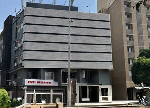 Um hotel de vários andares com linhas horizontais na fachada, uma entrada visível e uma placa com a inscrição 'HOTEL NEELKANE'.