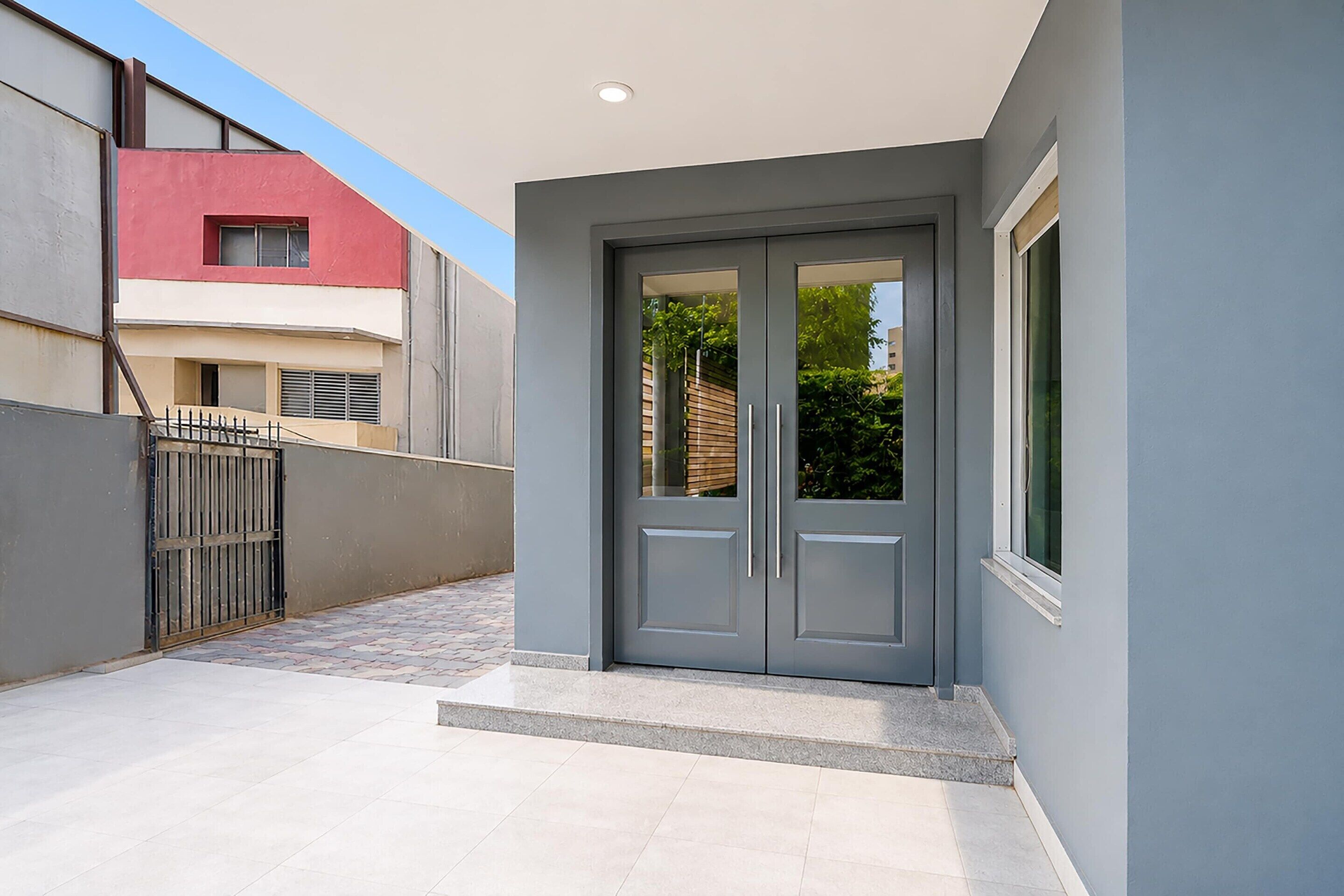 Un edificio moderno con una puerta gris, un ventanal pequeño, una verja metálica y un camino pavimentado.