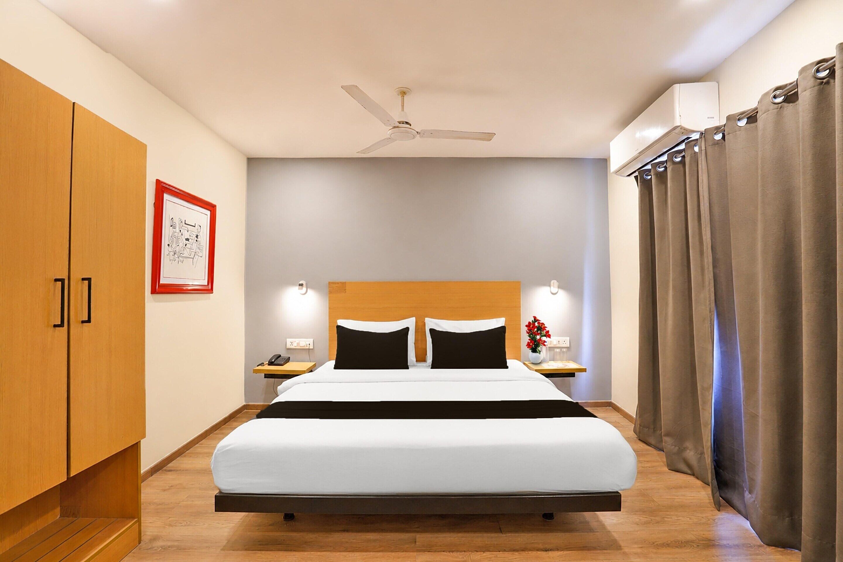 Habitación de hotel con cama, mesita de noche, teléfono, un cuadro en la pared, ventilador de techo y ventana con cortinas.