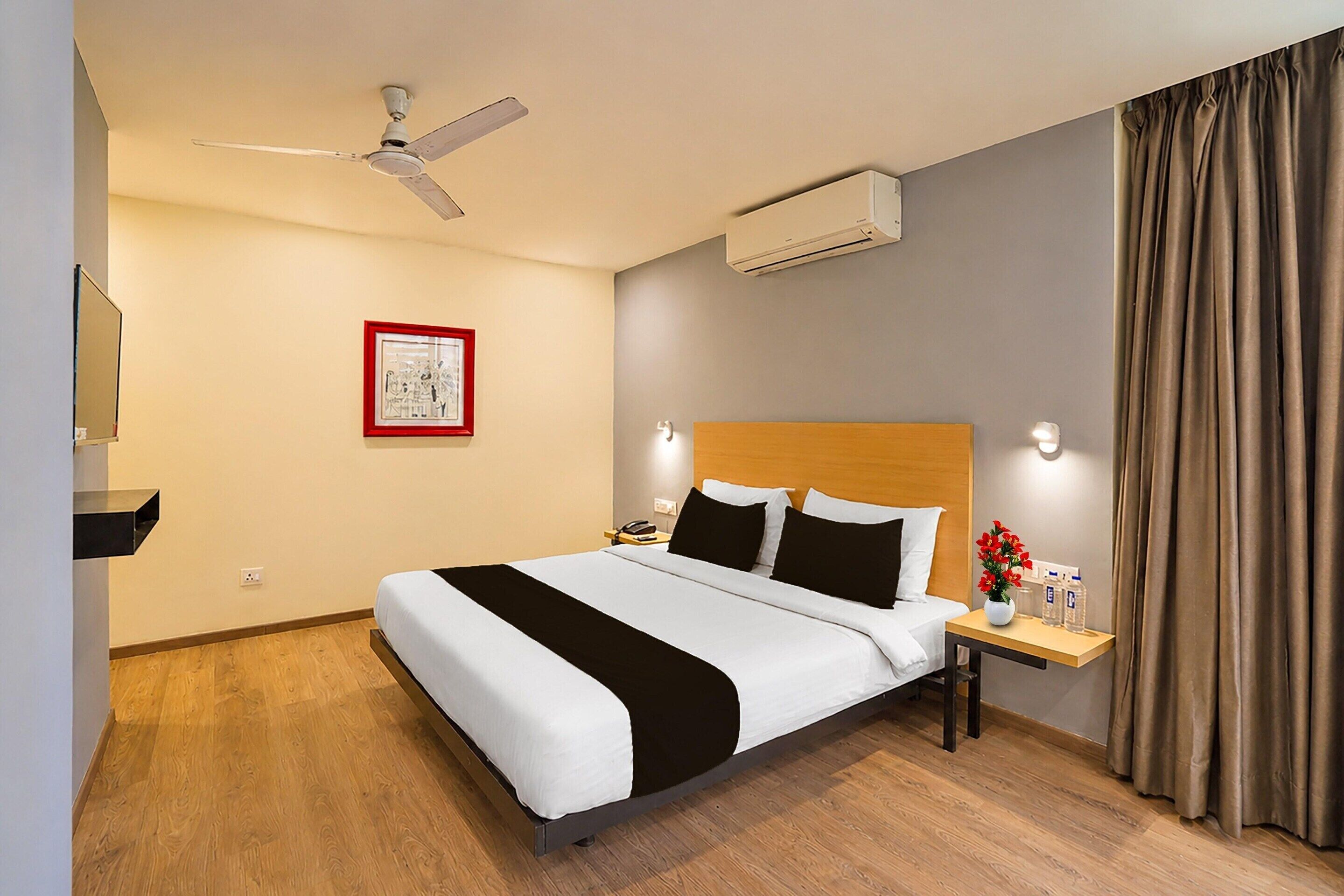 A hotel room with a bed, a nightstand, a ceiling fan, an air conditioning unit, a framed picture on the wall, and a window with curtains.