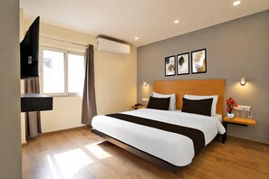 A hotel room with a bed, a bedside table with a lamp, a TV, and an air conditioning unit.