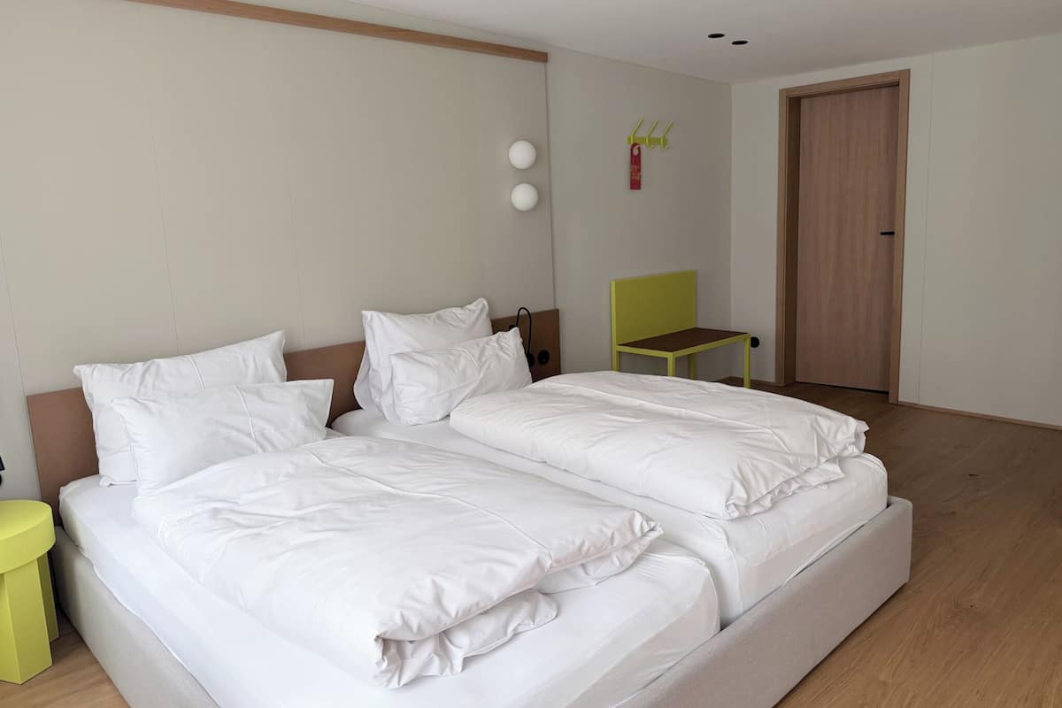 Ein Doppelbett mit weißer Bettwäsche, ein kleiner gelber Nachttisch, ein grüner Sessel, ein Holzschrank und eine Wandleuchte.