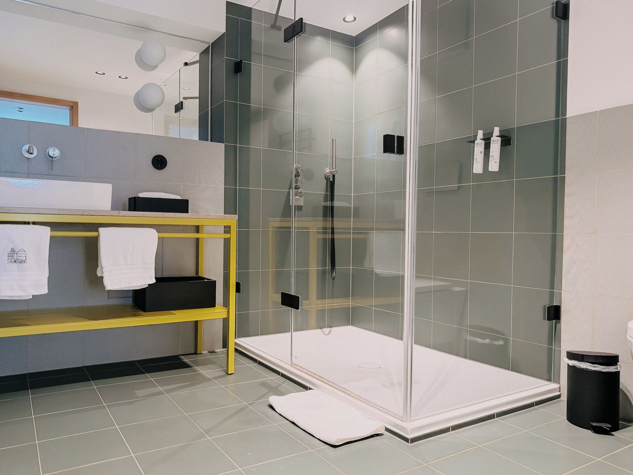 A modern bathroom with a glass-enclosed shower, a yellow shelf with towels, and a white sink.