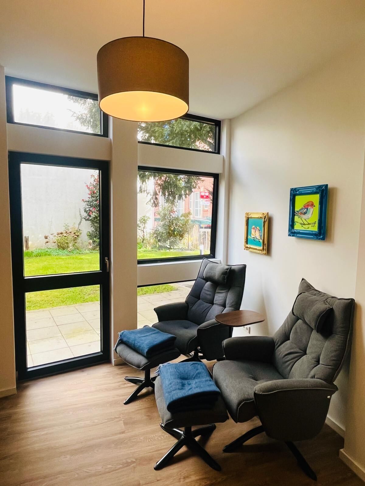 Een moderne woonkamer met een bank, poef en een tafel. Grote ramen bieden uitzicht op een tuin en gebouwen.