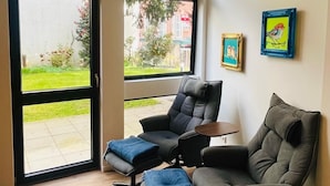 A modern living room with a sofa, ottoman, and a table. There are large windows with a view of a garden and buildings outside. - Exceptional apartment in a historic building with a view of the garden. (Bad Iburg)