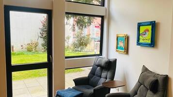 Een moderne woonkamer met een bank, poef en een tafel. Grote ramen bieden uitzicht op een tuin en gebouwen.