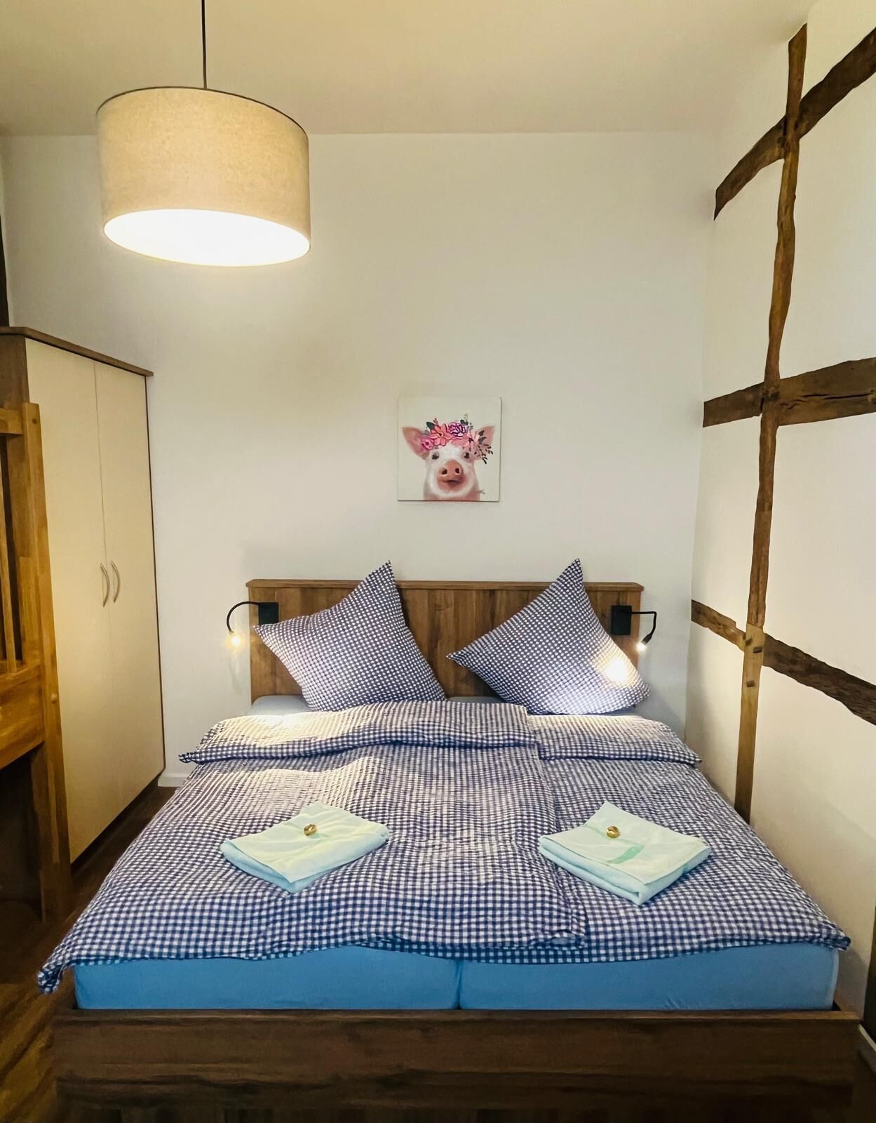 A bedroom with a bed, a hanging light, a wooden headboard, a wall-mounted picture, and a wooden wardrobe.