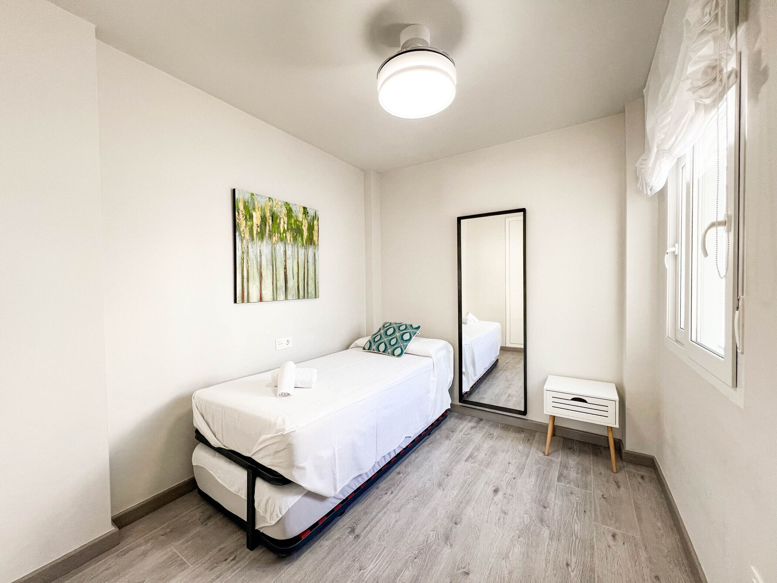 A single bed with a white mattress and black frame, a small bedside table with a white lamp, a mirror on the wall, and a window with curtains.