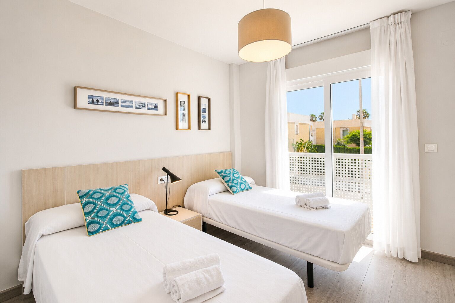 Habitació d'hotel amb dos llits, una tauleta de nit, una làmpada, una finestra amb cortines i vista al balcó.