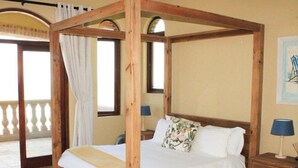 Ein Bett mit Baldachin, dessen hölzerner Rahmen einen weißen Überwurf und ein floraleres Kopfkissen trägt. Ein Nachttisch mit Lampe und ein Bilderrahmen an der Wand sind ebenfalls zu sehen.