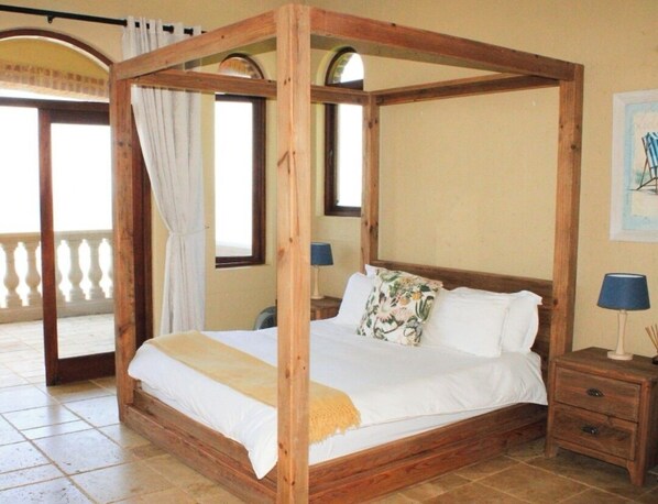 Een hemelbed met houten frame, een witte spreidek en een bloemenkussen. Er staat een nachtkastje bij met een lamp en er hangt een fotolijst aan de muur.