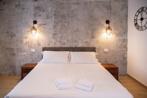 A neatly made bed with white linens and towels, two wall-mounted light fixtures, a wooden nightstand, and a clock on the wall.