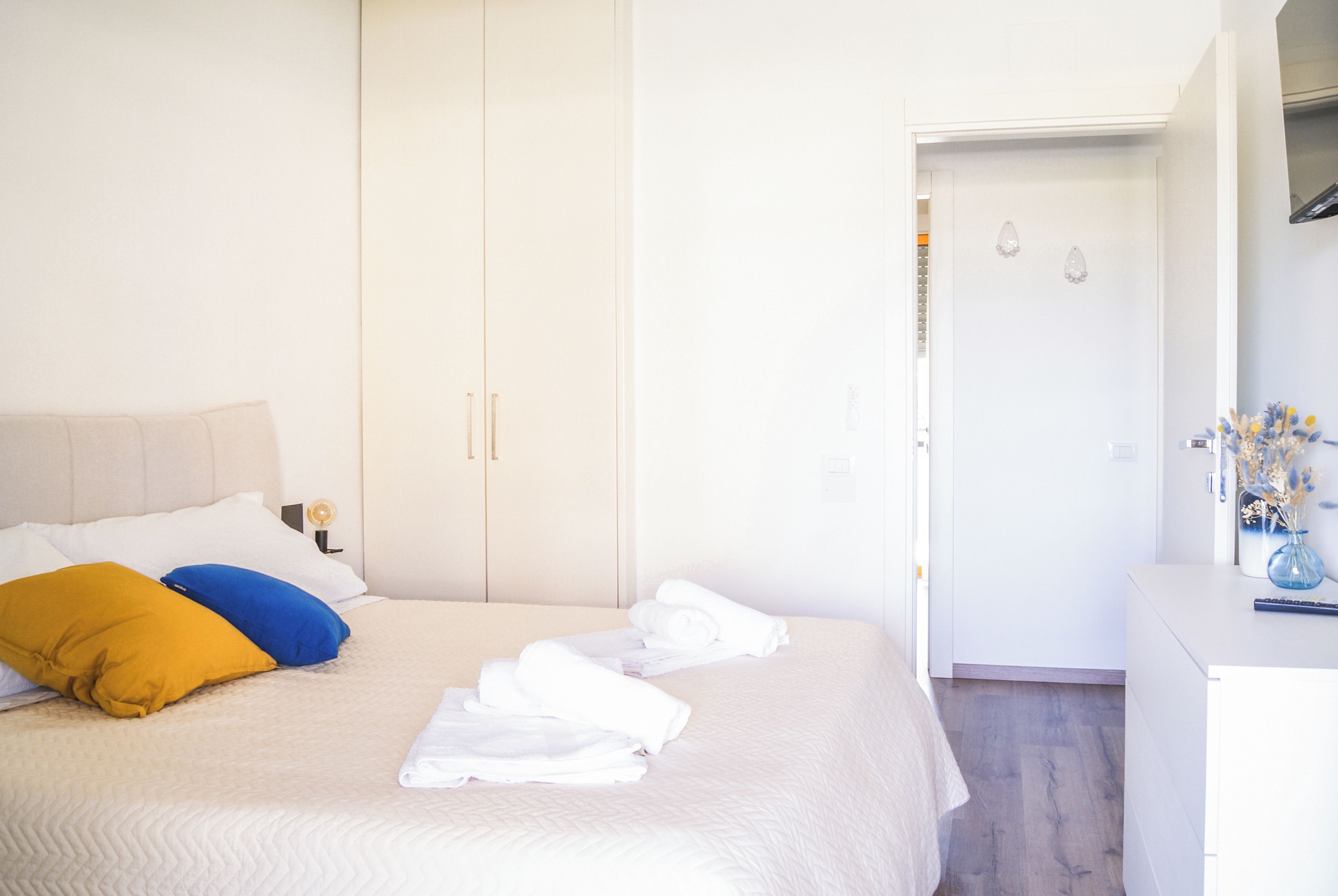 Un dormitorio con cama, armario, mesita de noche, lámpara y un jarrón con flores secas.