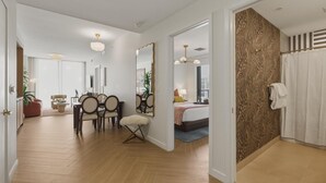 Moderni hotellihuone, jossa on puulattia, ruokailutila tuoleineen ja pöytineen, suuri peili, kuviollisella seinällä varustettu sänky ja suihkutila.
