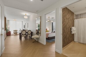 Ein modernes Hotelzimmer mit Holzboden, einem Essbereich mit Stühlen und Tisch, einem großen Spiegel, einem Bett mit gemustertem Wandhintergrund und einer Duschkabine.
