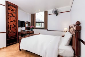 Ein Hotelzimmer mit einem hölzernen Bett, einem Fernseher, einem Fenster mit Vorhängen, einem Schreibtisch mit Lampe und einem dekorativen Raumteiler.