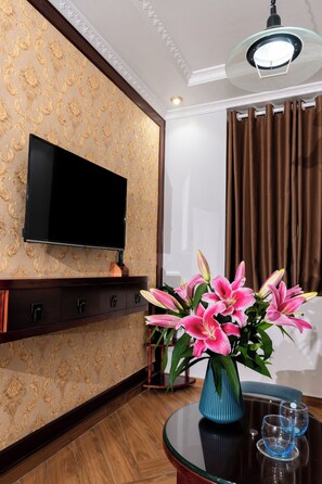 A room with a flat-screen TV mounted on a patterned wall, a dark wooden cabinet below, a round glass table with a vase of pink flowers, and a brown curtain. - The Eastern Hotel CatBi Airport (Hai Phong)