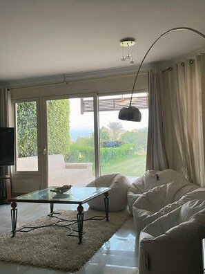 A modern living room with a glass coffee table, white sofa, and a view of a garden and mountains.