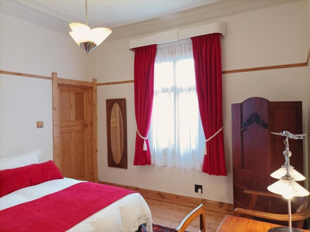 Een slaapkamer met een bed, een houten garderobe, een bureau met een lamp en een raam met rode gordijnen.