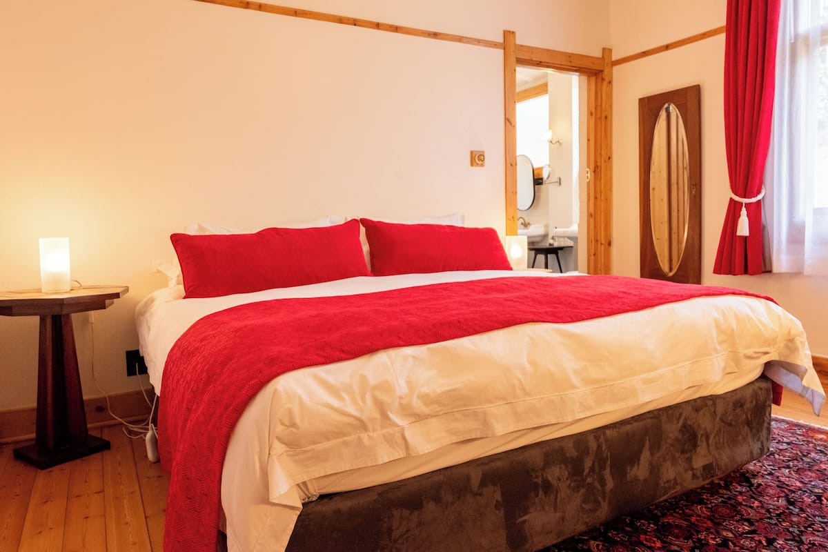 A bedroom with a bed, a bedside table with a lamp, a mirror, and a window with curtains.