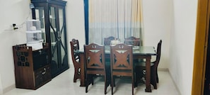 A dining area with a glass-top table, wooden chairs, a cabinet, and a window with curtains.