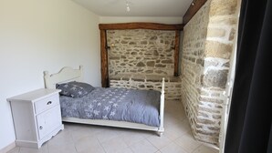 A bedroom with a stone wall, a bed with a grey star-patterned bedspread, a white bedside table, and a wooden ceiling beam. - Gite l'écurie, in a restored farmhouse, fully equipped with pellet stove (Surtainville)