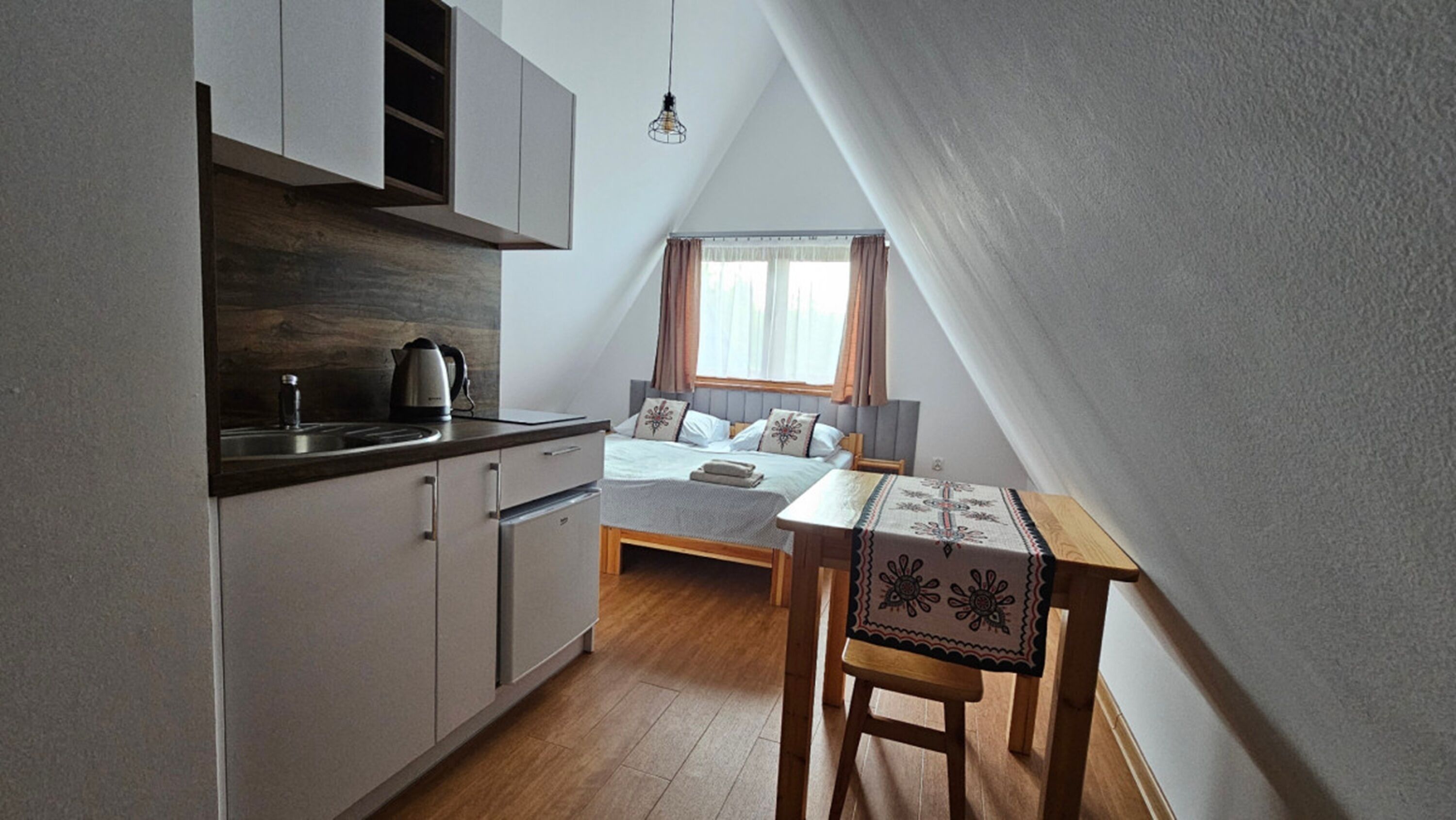 Een kleine, moderne kamer met een kitchenette, een bed met hoofdeinde, een raam met gordijnen, en een tafel met een tafelkleed met dessin.