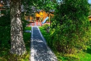 Un sendero conduce a un parque infantil con un tobogán, rodeado de árboles y césped.