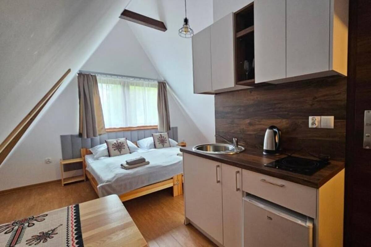 Ein kleines Schlafzimmer mit Bett, Nachttisch und einem fensterbelaubten Fenster. Daneben eine Küchenzeile mit Spüle, Herd und Schränken.