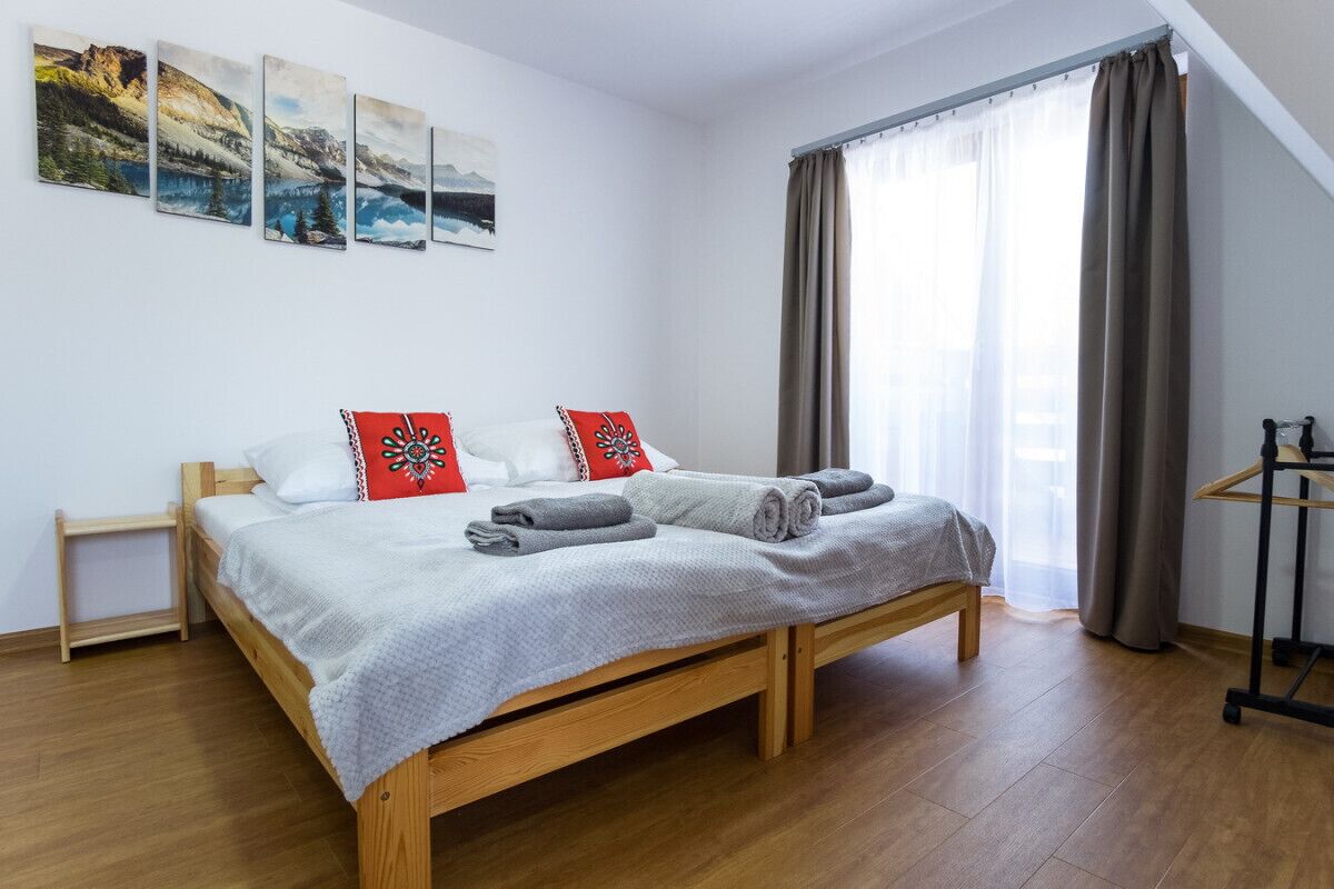 Un dormitorio con cama de madera, ropa de cama blanca, almohadas rojas y blancas, una mesita de noche pequeña y una ventana con cortinas.