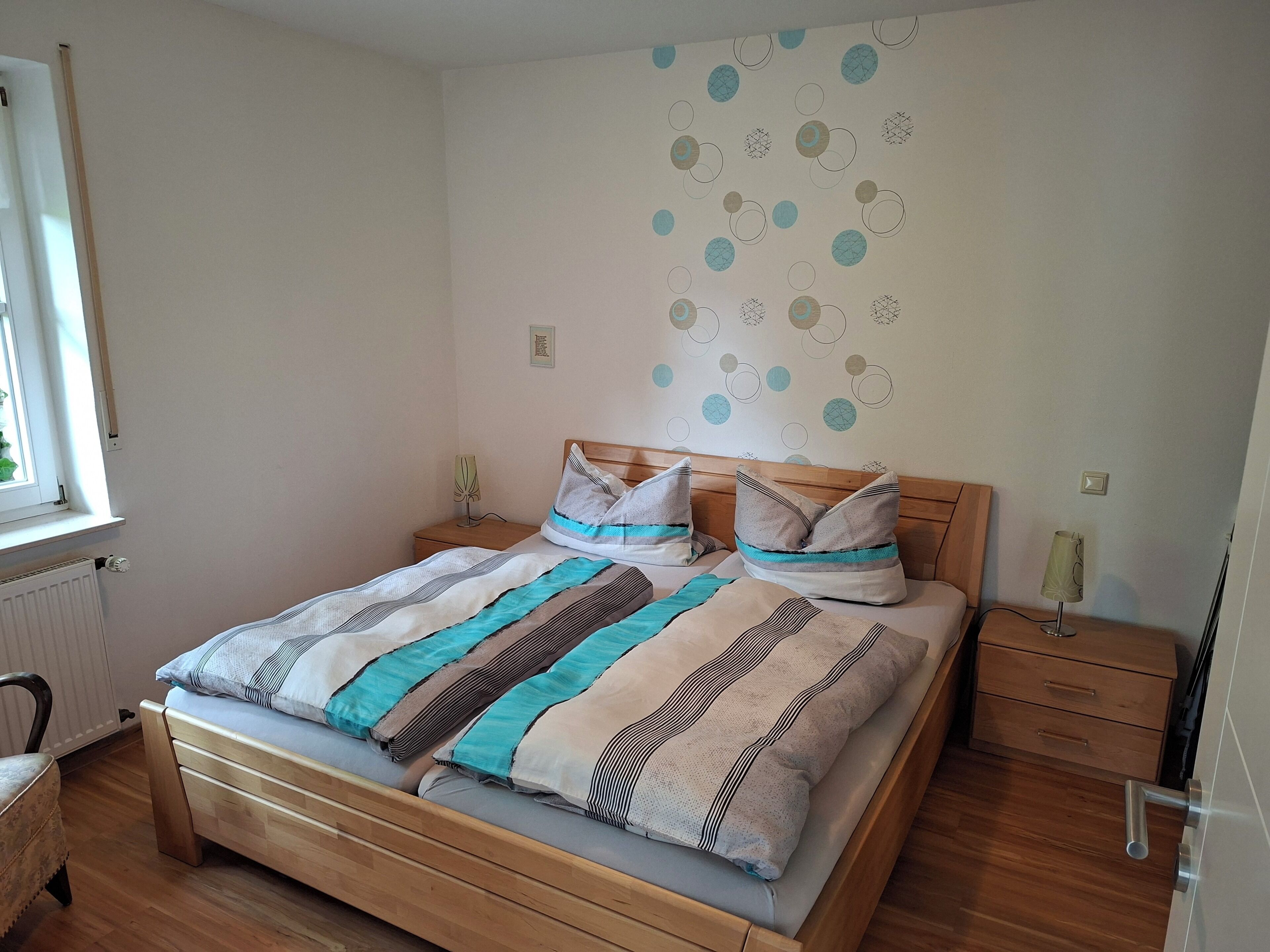 A neatly made bed with striped bedding, a wooden headboard, and a bedside table with a lamp.. Vacation apartment in Marsberg (Sauerland) with terrace directly on the Diemel cycle path