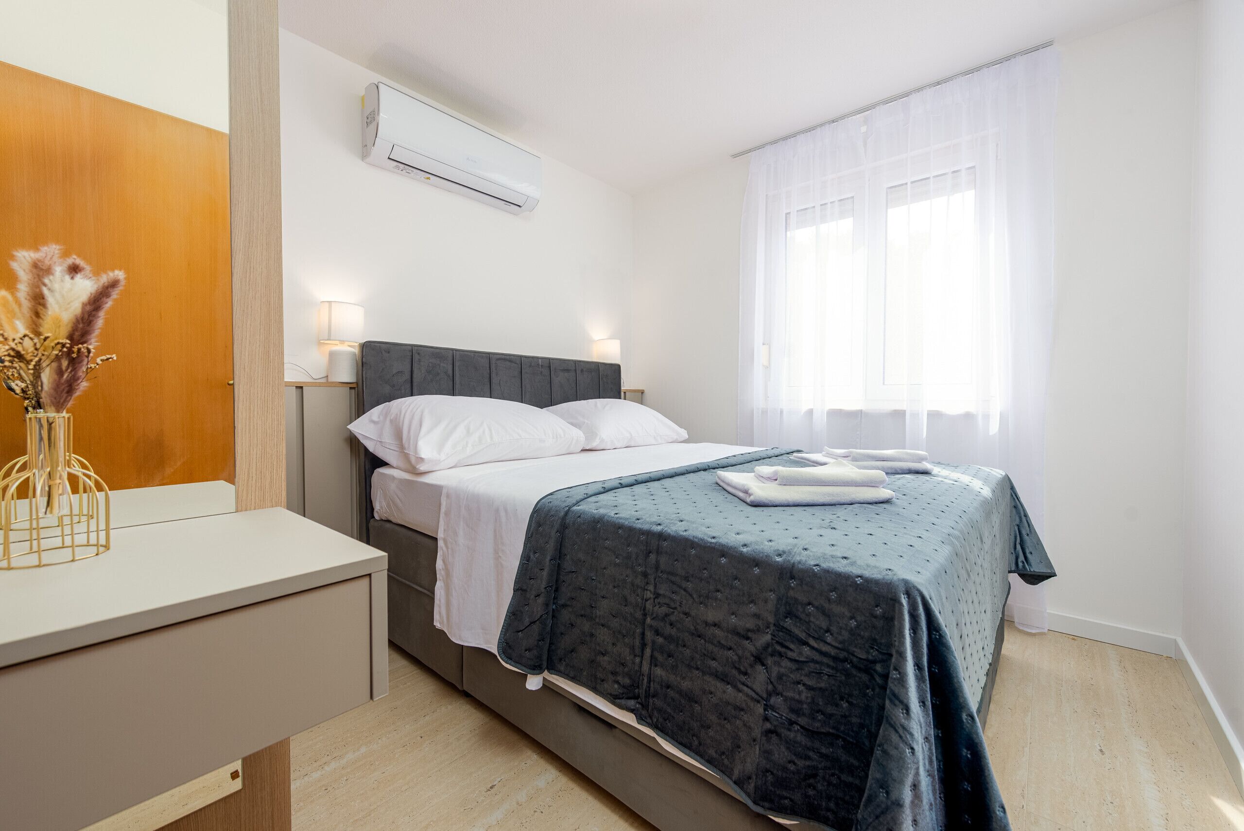 A bedroom with a bed, bedside table, lamp, air conditioning unit, and a window with curtains.