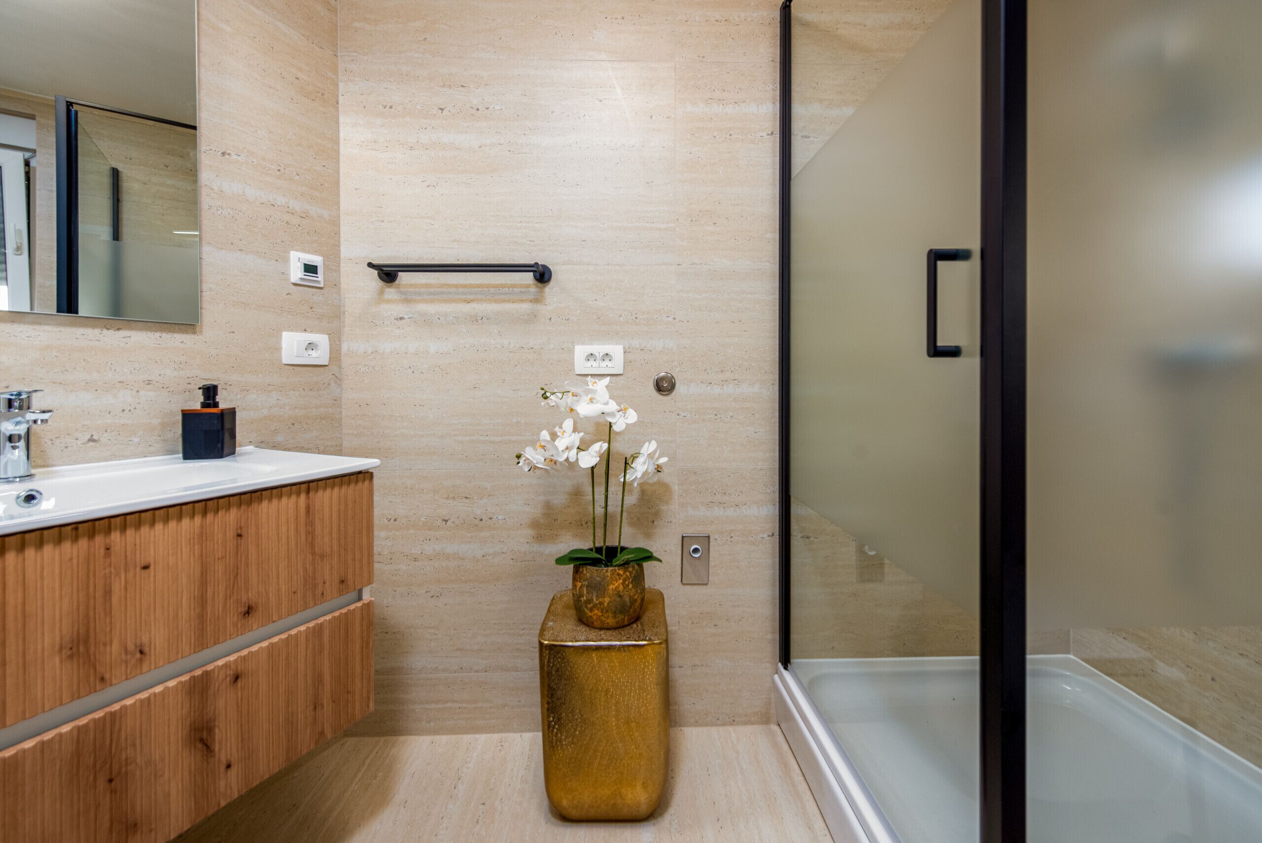 A modern bathroom with a wooden vanity, a glass shower enclosure, and a vase with white flowers.