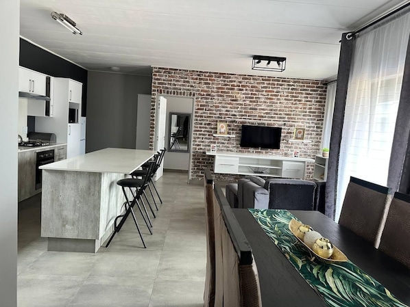 Eine moderne Küche mit einer zentralen Kochinsel, einem Essbereich mit Fernseher und einer Wand mit Backsteinverkleidung.